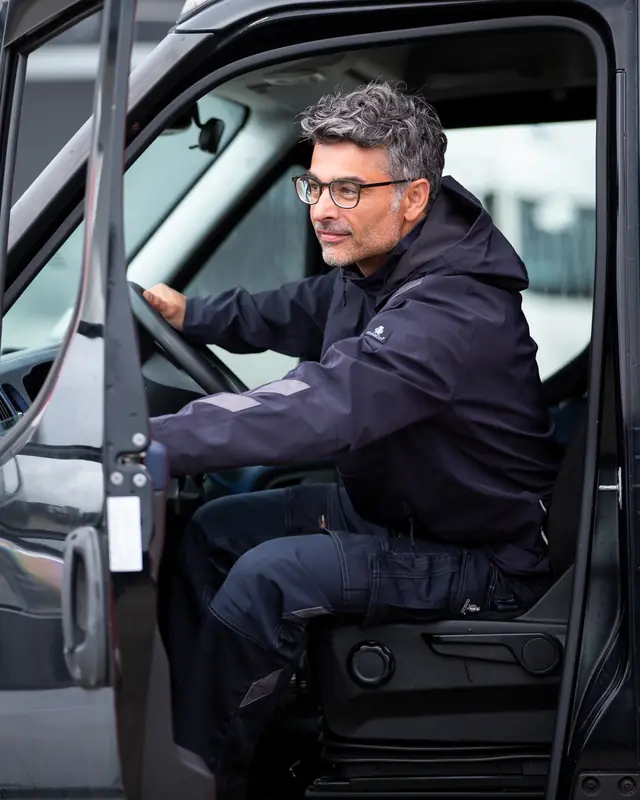 Mann in dunkelblauer MASCOT UNIQUE Hardshell-Jacke am Steuer eines Transporters